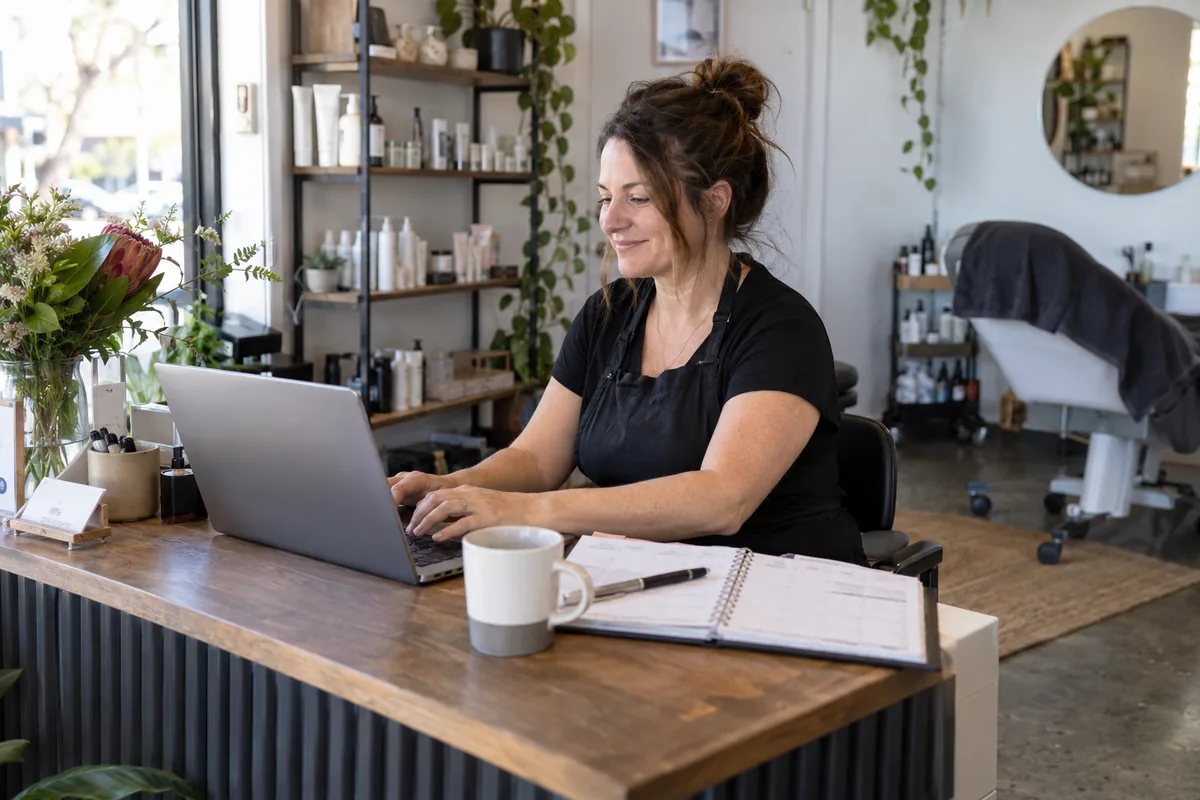 A salon owner uses a laptop at reception with an appointment diary beside her.